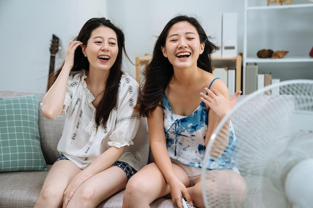 two happy smiling young asian female friends looking at electric fan with cold breeze wind blowing on hair sitting on couch sofa in living room. girls relax with air cooler at home on summer break.