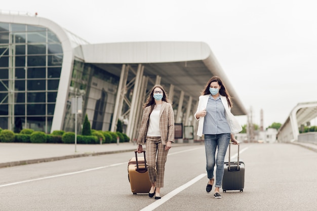 Two happy women in protective masks after coronavirus quarantine with suitcases go to the airport.