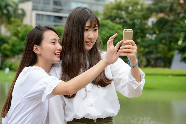 Two happy young beautiful Asian teenage women taking selfie together at the park