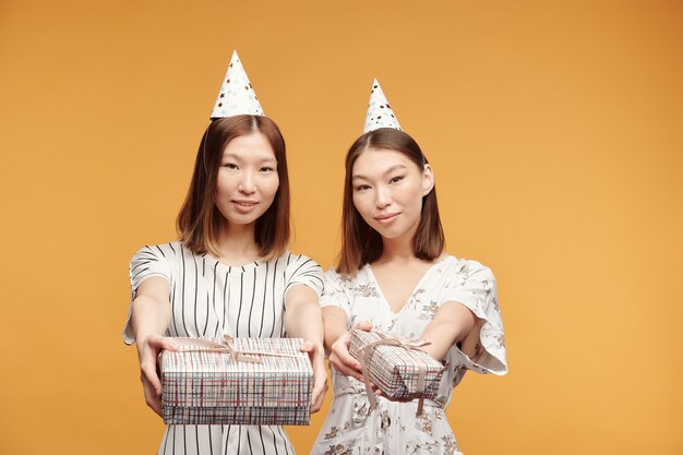 Two happy young beautiful female twins in smart dresses and birthday caps looking at you while passing packed giftboxes with presents