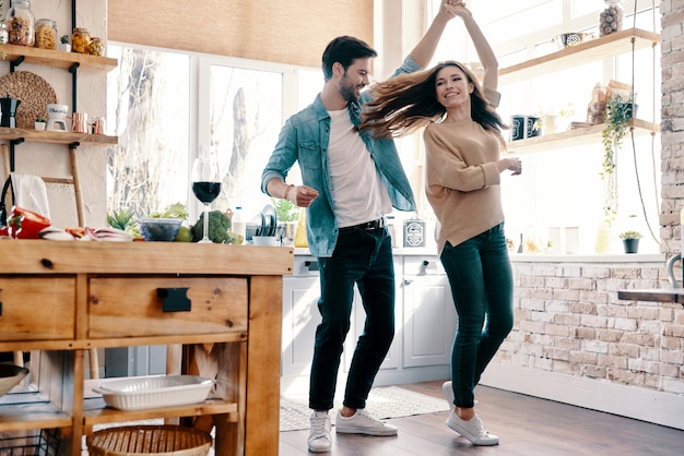 Two hearts filled with love. Full length of beautiful young couple in casual clothing dancing and smiling while standing in the kitchen at home