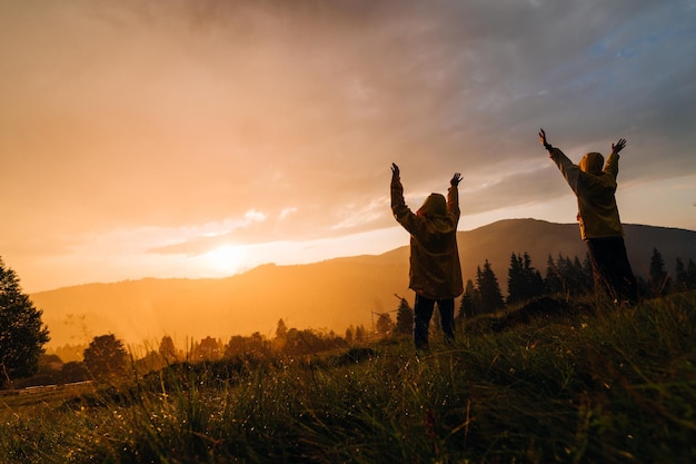 Two hikers stand in the rain on a meadow in the mountains and watch the incredible orange sunset