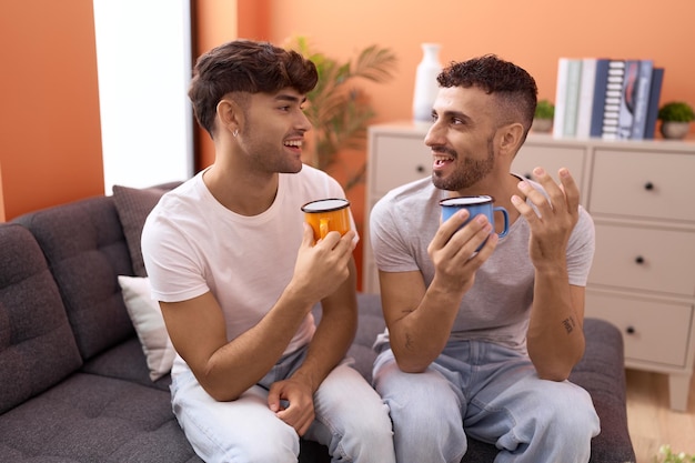 Two hispanic men couple drinking coffee sitting on sofa at home