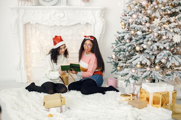 Two indian girl in eyeglasses sitting near Christmas tree and reading a book