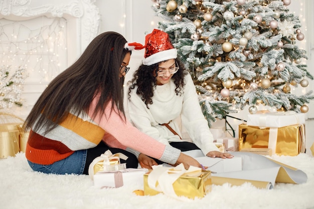 Two indian girl in eyeglasses wrapping a gift in a paper near Christmas tree