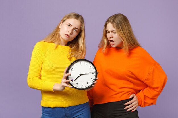 Two irritated young blonde twins sisters girls in vivid colorful clothes hold round clock isolated on pastel violet blue wall. People family lifestyle concept. 