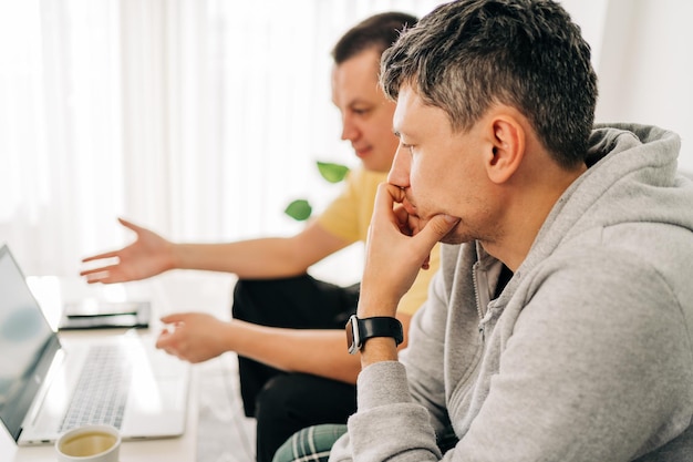 Two it freelancer men typing in laptop working from home office two programmers sitting on couch