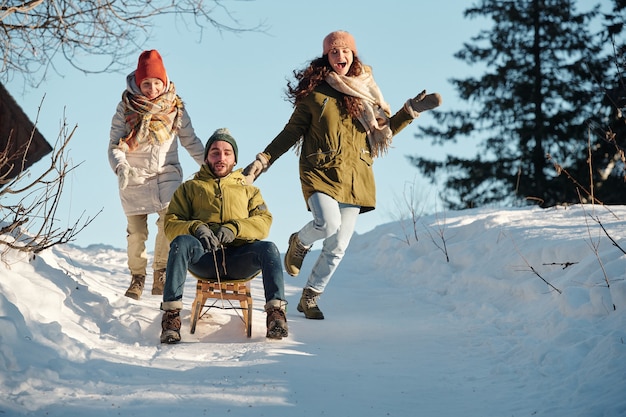 Two joyful girls in winterwear running after young man on sledge moving down hill among snowdrifts in the forest while having fun on winter day