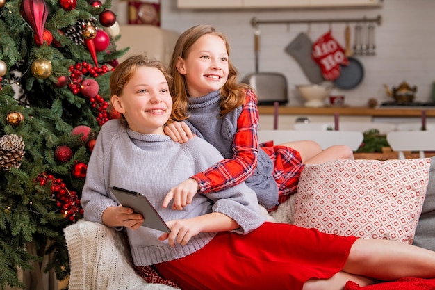 Two joyful sisters are sitting at home near the christmas tree and looking at tablet monitor