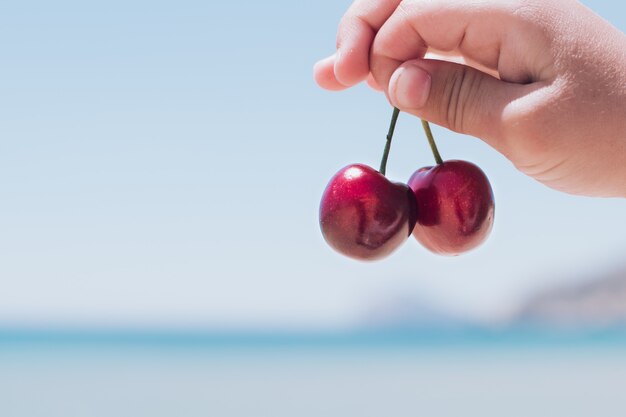 Two juicy cherry berries in hand of the child against the blue sea and sky.