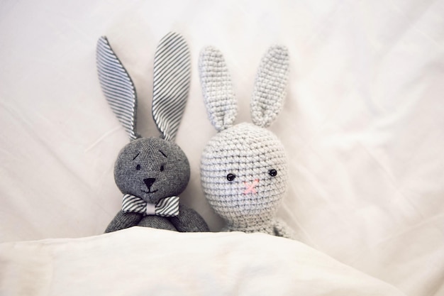 Two knitted rabbit toys lie under a blanket in a white bed