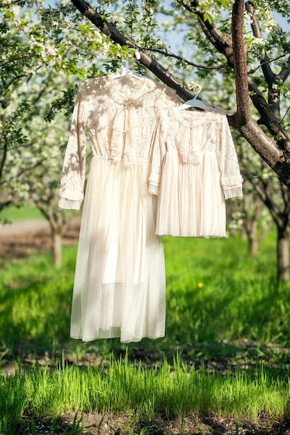 Two lace beige dresses for mom and daughter hang on branches in a blooming apple orchard