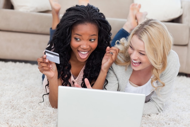 Two laughing women are lying on the floor with a laptop and a bank card