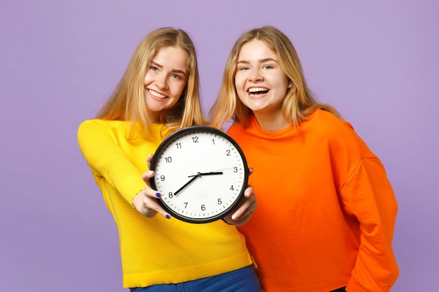 Two laughing young blonde twins sisters girls in vivid colorful clothes hold round clock isolated on pastel violet blue wall. People family lifestyle concept.