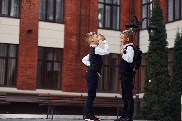 Two little boys in school uniform that is outdoors together give high five near education building.