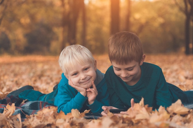 Two little brothers using smartphone lying in yellow autumn leaves Smiling and having fun Fall day