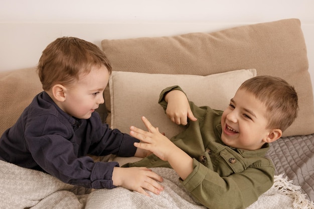 Two little and cute caucasian boys playing together on the bed at home. Interior and clothes in natural earth colors. Cozy environment. Children having fun, two brothers kittle each other.