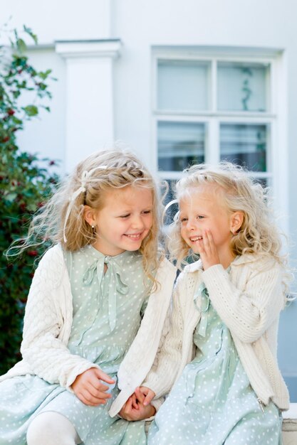 Two little cute girls are sitting in the garden and sharing secrets One girl whispers something in another's ear The two sisters have a close relationship