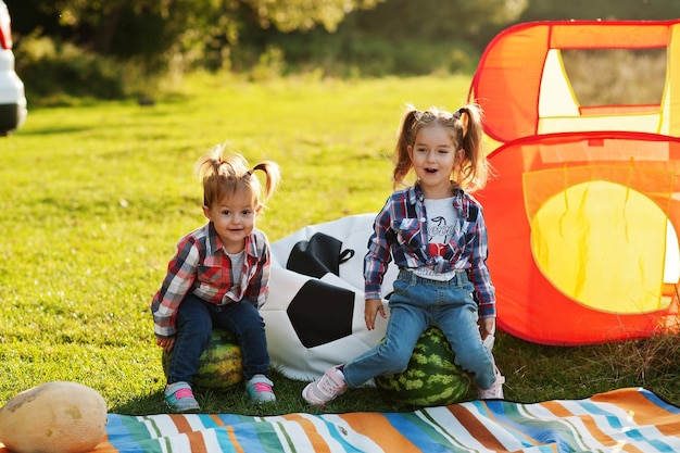 Two little cutie sister girls outdoor in picnic wear checkered shirt Sitting on watermelon