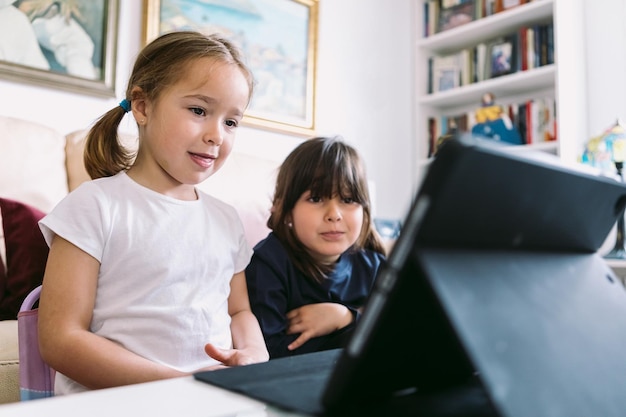 Two little girls attentively watching a video on a tablet in the living room at home and smiling Concept of childhood technology learning having fun the Internet and connectivity