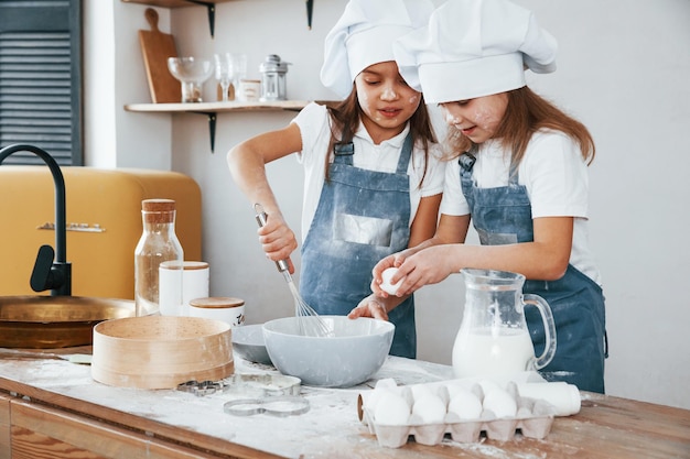 Two little girls in blue chef uniform mixing food on the kitchen by using flour and eggs