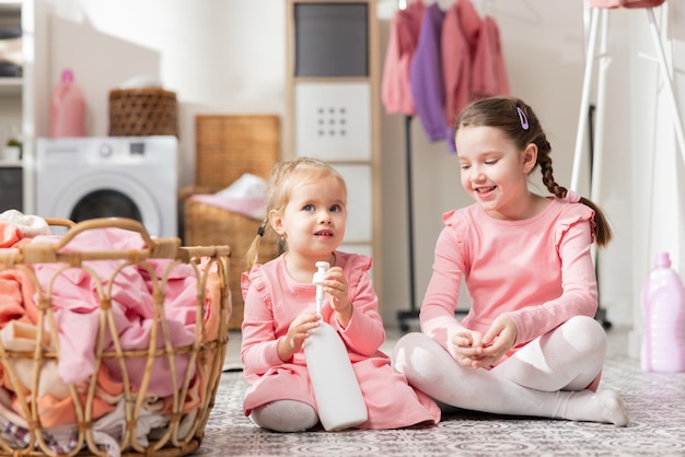 Two little girls in pink dresses sitting on the laundry room floor by a basket of clothes