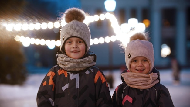 Two little kids standing on public ice rink and waving with their hands  looking in the camera