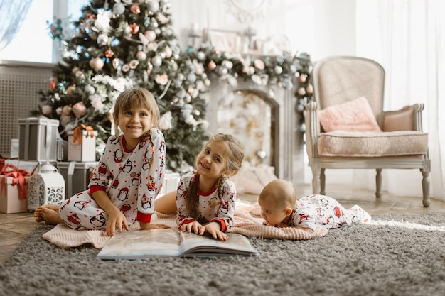 Two little sisters and a tiny brother lie on the carpet and read book near the New Year's tree