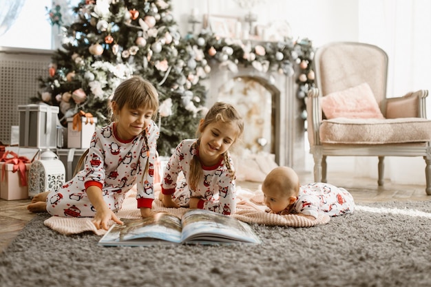 Two little sisters and a tiny brother lie on the carpet and read book near the New Year's tree