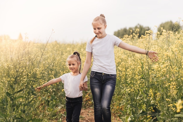 Two little sisters walk around the flowering canola in summer. two little sisters hold hands, stand in a field.