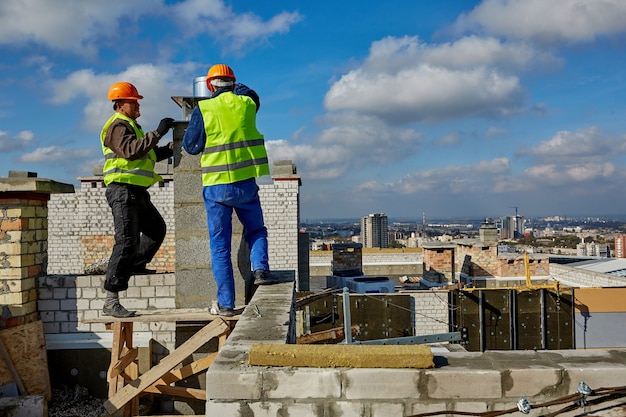 Two males professional workers in protective clothing and hardhats are working with ventilation system on roof of building under construction