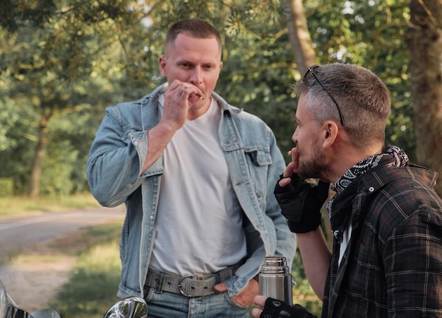 Two man friends of motorcyclists bikers on motorcycles smoking on the side of the road and talking