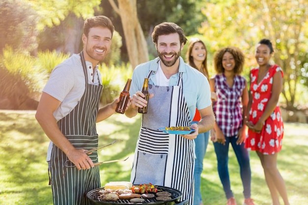 Two men holding a beer bottle while preparing barbecue grill