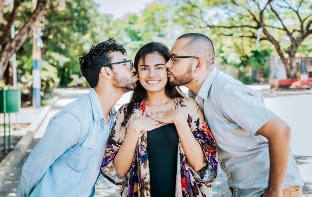 Two men kissing a girl cheek Portrait of two guys kissing a girl cheek Two young men kissing a woman cheek outdoor love triangle concept Polygamy concept