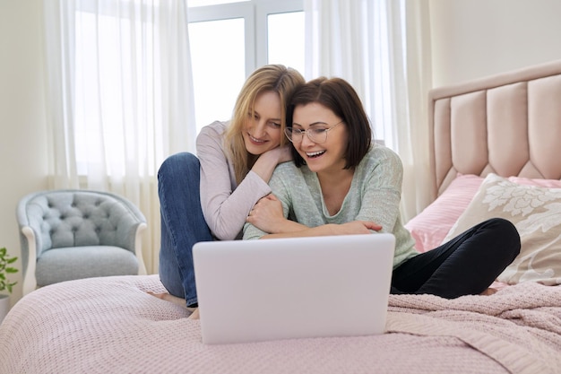 Two middle aged women having rest sitting together at home on bed looking in laptop screen