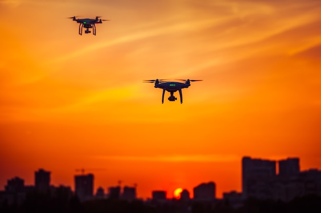 Two modern remote control air drones fly with action cameras in dramatic orange sunset sky cityscape