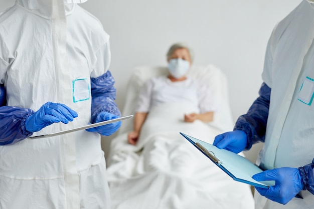 Two modern unrecognizable doctors wearing protective clothes standing in hospital ward with senior female patient on bed discussing something