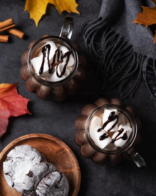 Two mugs of aroma hot chocolate with marshmallows on a dark background with autumn maple leaves cookies and gray scarf Concept cozy drink Top view