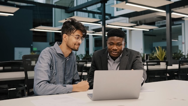 Two multicultural professional male coworkers men colleagues at office use laptop discuss project