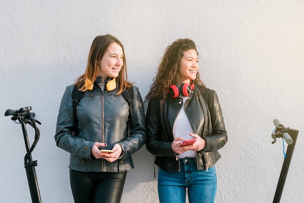 Two multiethnic female friends on electric scooters using smartphone outdoors