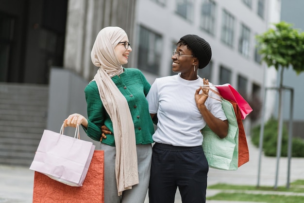 Two multiethnical girls walking down the old city street after shopping carrying colorful bags