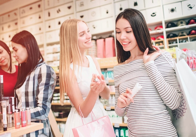 Two nice girls in the front are standing and smiling. Asian girl is looking at cosmetics that blinde girl has in her hand. Blonde girl is looking to asian girl and smiling