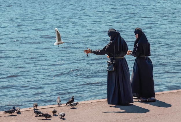 Two nuns feed birds on the river embankment