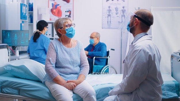 Two old senior persons at doctor wearing masks and talking with nurse and doctor in protective wear. Modern private clinic or hospital ward during COVID 19 pandemic. Healthcare medicine appointment