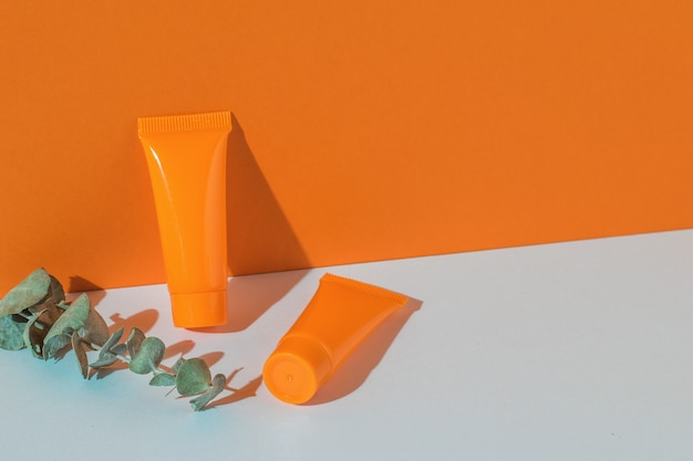 Two orange tubes of cream and a sprig of eucalyptus on an orange and white background. Place for text.