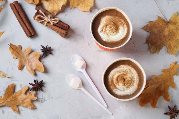 Two paper cups with tasty pumpkin latte and spice on gray table with autumn leaves. Top view. Closeup.