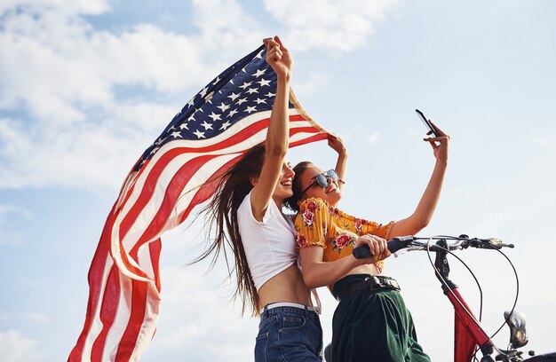 Two patriotic cheerful women with bike and USA flag in hands makes selfie.