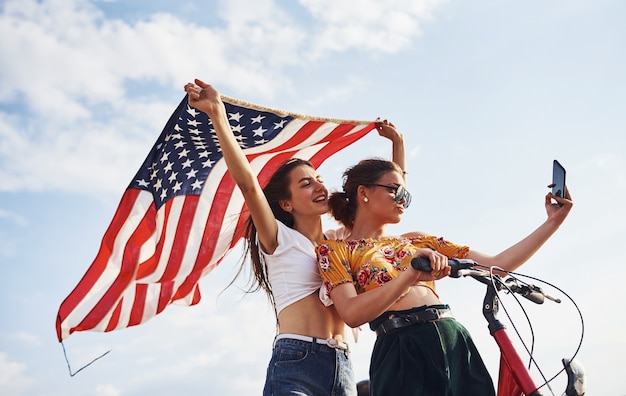 Two patriotic cheerful women with bike and USA flag in hands makes selfie.