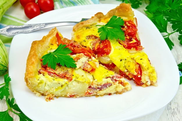 Two pieces pie of zucchini with tomatoes and eggs in a white plate on a napkin, parsley on the background light wooden boards