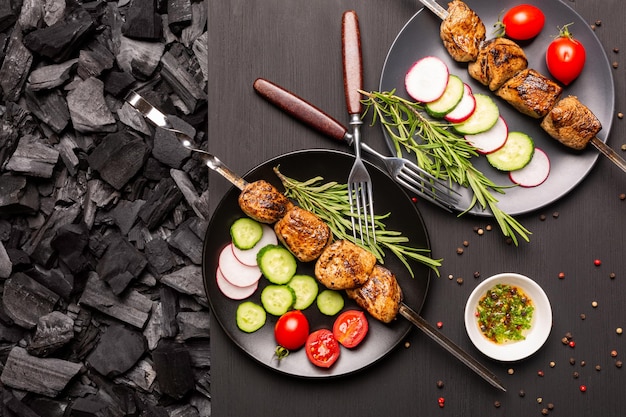 Two plate with kebab and fresh vegetables on a black wooden tabletop and charcoal  for barbecue. Top view.